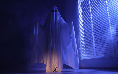 Creepy ghost. Woman covered with sheet near window in blue light, low angle view