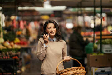 A happy woman with basket is using her phone at farmers market.