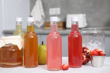 Tasty kombucha in glass bottles, jar and fresh fruits on white tiled table