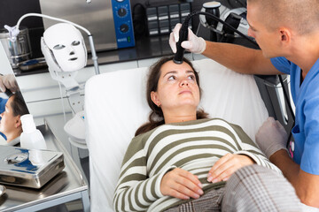 Naklejka premium Young woman getting facial electroporation procedure from experienced male cosmetologist in aesthetic medicine office