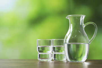 Jug and glasses with clear water on wooden table against blurred green background. Space for text