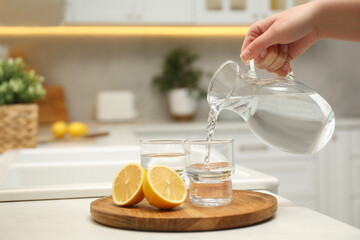 Woman pouring water from jug into glass at white table in kitchen, closeup