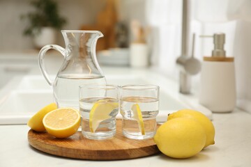 Jug, glasses with clear water and lemons on white table in kitchen
