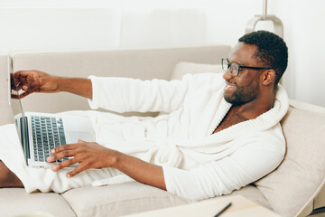 Smiling African American Freelancer Working on Laptop in Modern Home Office with a view of the Cityscape The young black guy, dressed in a bathrobe, is sitting on a comfortable sofa, typing on his