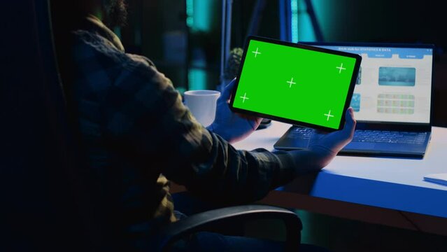 Man in home office checking statistical financial graphs on laptop, crosschecking with data on mockup tablet. Remote employee holding green screen device, analyzing business graphics