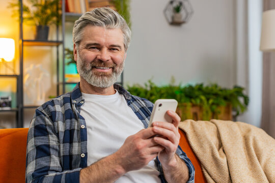 Happy mature man sitting on sofa at home getting message from old friend using smartphone look aside with smile. Caucasian smiling old man reading good news on phone screen in living room at home - Powered by Adobe