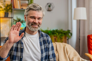 Caucasian mature man smiling friendly at camera and waving hands gesturing hello hi greeting welcoming with hospitable expression at home. Senior guy sitting on sofa in living room at home. Copy-space