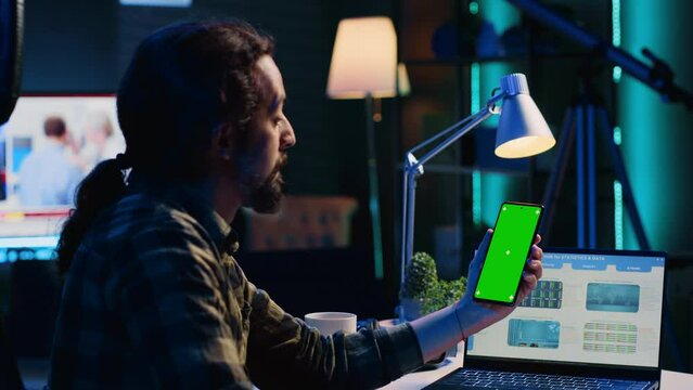 Teleworker holding green screen smartphone, analyzing statistical charts on laptop in living room. Self employed man checking business revenue numbers on mockup mobile phone