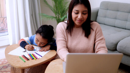 Latin American mother taking care of her child painting while using computer laptop at home. Family...