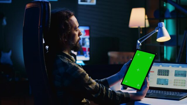 Remotely working man in home office checks statistical financial graphs on laptop, crosschecking with data on chroma key tablet. Freelancer holding isolated screen device, analyzing business graphics