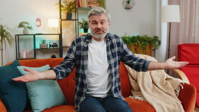 I don't know what to say. Confused senior bearded man feeling embarrassed about ambiguous question having doubts no idea being clueless and uncertain at home. Mature guy sits on sofa in living room.