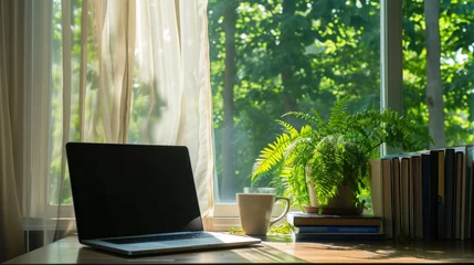 Fototapeten A modern laptop computer sits on a sleek desk next to a large window, offering a view of a lush green landscape. A cup of coffee and a fern plant sit beside the laptop © Vodkaz