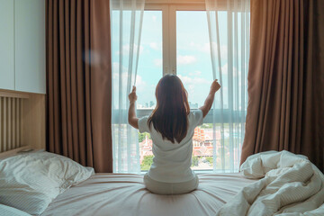 happy woman stretching on bed after wake up, young adult female rising arms and looking to window...