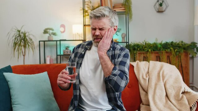 Senior bearded man feel terrible toothache after drink cold water. Middle aged guy sitting on sofa in living room touching cheek, feel hurt and suffering from sensitive tooth ache, pain and cavities.
