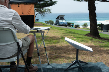 man with satellite internet dish connected working on computer at remote beach side campsite