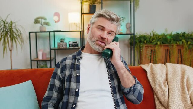 Sad senior man with gray hair talking on vintage retro wired telephone at home apartment. Disinterested Caucasian guy having annoyed boring conversation with hotline helpline service sitting on sofa.