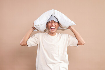 Young Asian man with eye mask putting a pillow on his head and screaming, having trouble sleeping