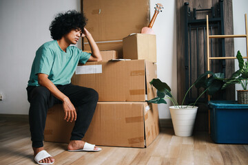Asian man with curly hair sitting near pile of carboard boxes showing confused and sad expression