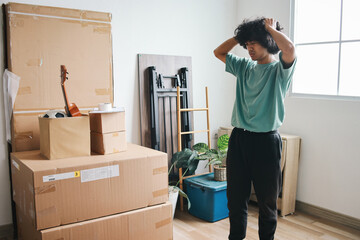 Exhausted young Asian man with curly hair touching heads, working hard after moving to new apartment, unpacking stuff