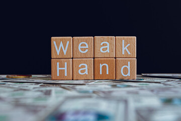 Wooden blocks with the text "Weak Hand" on a black background and crypto banknotes scattered on the ground.