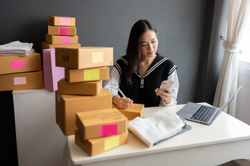 Asian female business owner working diligently To make the business grow In the office, working with laptops and smartphones, there are boxes of products ready to be sent to customers.