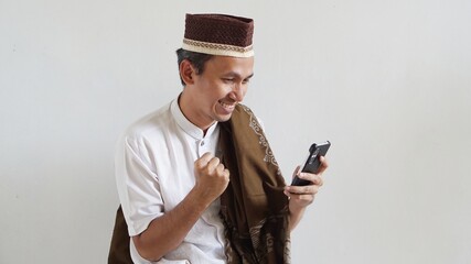 Fototapeta premium asian man with happy expression, playing mobile games, wearing cap and sarong