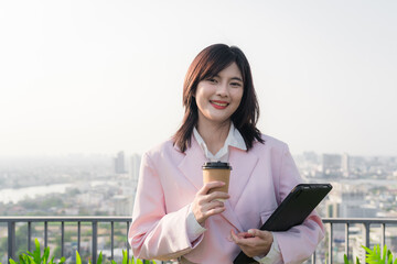 Beautiful young asian businesswoman using digital tablet and holding coffee on rooftop or skyscraper terrace in summer day morning, shopping online, concept of successful business development