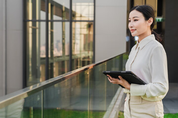 Asian woman using digital tablet look in distance planning or visualizing future success, dreaming or thinking, standing on rooftop at sunset. Business vision concept.