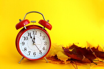 There is a red alarm clock with yellow autumn leaves on a yellow background.	