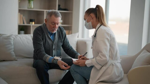 Therapist measure pressure senior man. Woman geriatric doctor in medical mask using tonometer checks pressure to elderly patient in modern clinic. Medical examination in hospital. Check up pensioner.