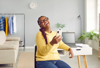 Happy cheerful joyful young African American woman in yellow sweater sitting at working desk with laptop computer at home, having break, holding mobile phone and laughing at funny joke on social media