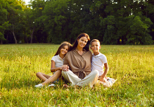 Happy family spending time in nature. Portrait of a mother and children in the park in summer. Young woman together with her kids sitting on fresh green grass, with beautiful trees in the background