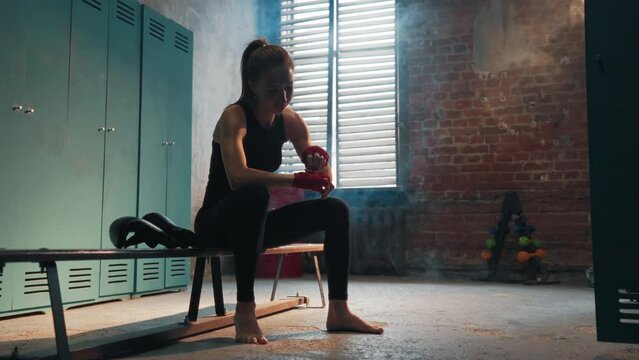 Focused young woman fighter boxer wraps hands wrists with boxing bandage sits on bench in locker room. Girl kickboxer prepares for battle fight. Professional sport, competition, preparation concept.