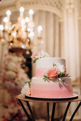 elegant white two-tiered wedding cake decorated with flowers on a small table against the backdrop of a wedding arch