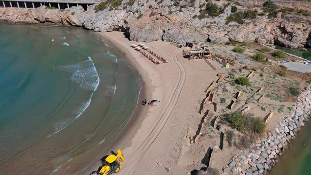 Yellow excavator working on idyllic beach near port ginesta beach in barcelona, spain with clear waters and sandy shore, aerial view