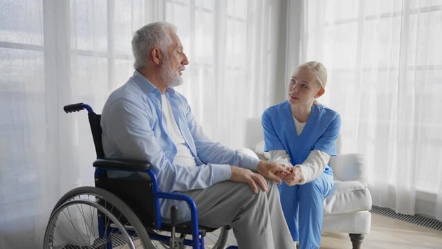 Nurse talking senior man in wheelchair. Female social welfare physical therapist looks after senior male, providing medical care at home. Living with legs paralysis, retirement, special needs people.
