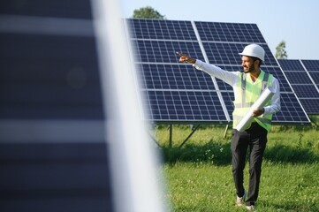 Portrait of Young indian man technician wearing white hard hat standing near solar panels against blue sky.Industrial worker solar system installation, renewable green energy generation concept.
