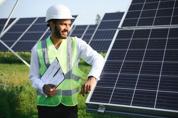 Portrait of Young indian man technician wearing white hard hat standing near solar panels against...