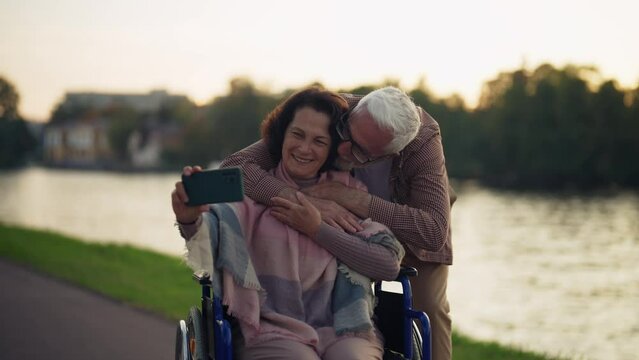 Wife in wheelchair makes couple selfie. Senior family spending time in city park near pond taking photos on smartphone. Man hugging embrace woman showing love care. Living with physical disability.