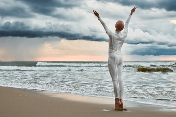 Hairless girl with alopecia in white futuristic suit standing on sea beach stretched out arms to cloudy sky, full length back view portrait, metaphoric performance with bald sensitive teenage girl - obrazy, fototapety, plakaty