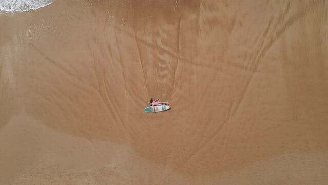 Aerial top view of slender woman in pink bikini lying on sandy shore with surfboard. Pretty surfer girl relax after surfing session on beach sand washed by ocean waves sea surf. Camera fly up.