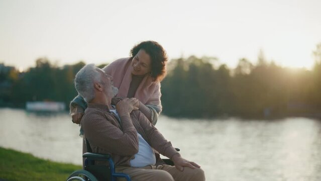 Wife walking with husband in wheelchair. Senior family couple spending time in city park near pond. Woman hugging supporting man showing love care, they talking. Live with physical disability concept.