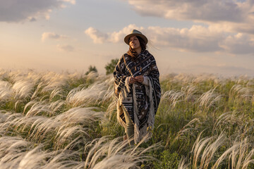 A woman in a hat and an Indian poncho on a feather grass field on a cloudy day, against a sky with clouds.