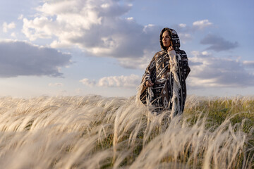 Woman in a Peruvian poncho, a hood on her head. Summer landscape, feather grass field, sky with clouds.
