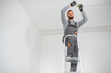 Repairman or professional electrician in workwear installing light spots, standing on the ladder in...