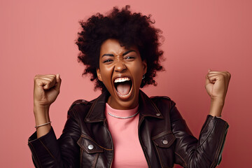 Excited contented curly haired African-American young woman wearing leather jacket is celebrating with her arms raised and huge smile on her face against pink background