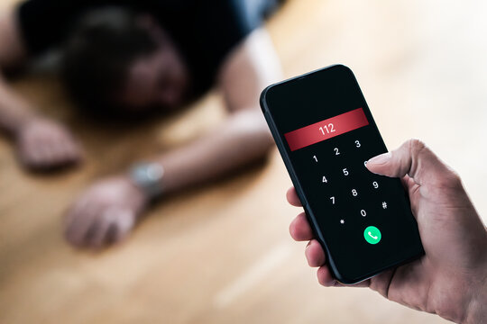 Emergency call 112. Accident or epilepsy. Unconscious fainted young man lying on the floor. SOS ambulance alarm with phone in hand. Urgent medical help aid. Alcohol poisoning or overdose at a party.