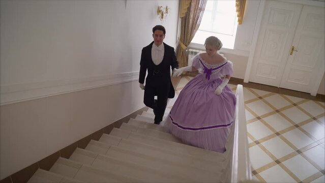 A couple in love in a 17th century image. An elegant couple, a girl in a beautiful dress and a guy in an 18th century look. A beautiful couple in 19th century costumes is walking up the stairs.
