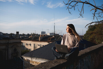 Woman freelancer uses laptop on cement wall outdoors against the sky and the roof of the city. The woman to be focused on her work or enjoying some leisure time while using her laptop.