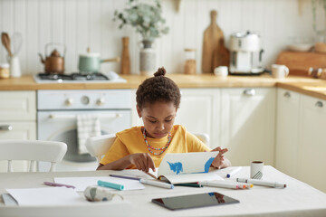 Gen Alpha African American girl sitting at table in cozy kitchen flipping pages of her sketchbook